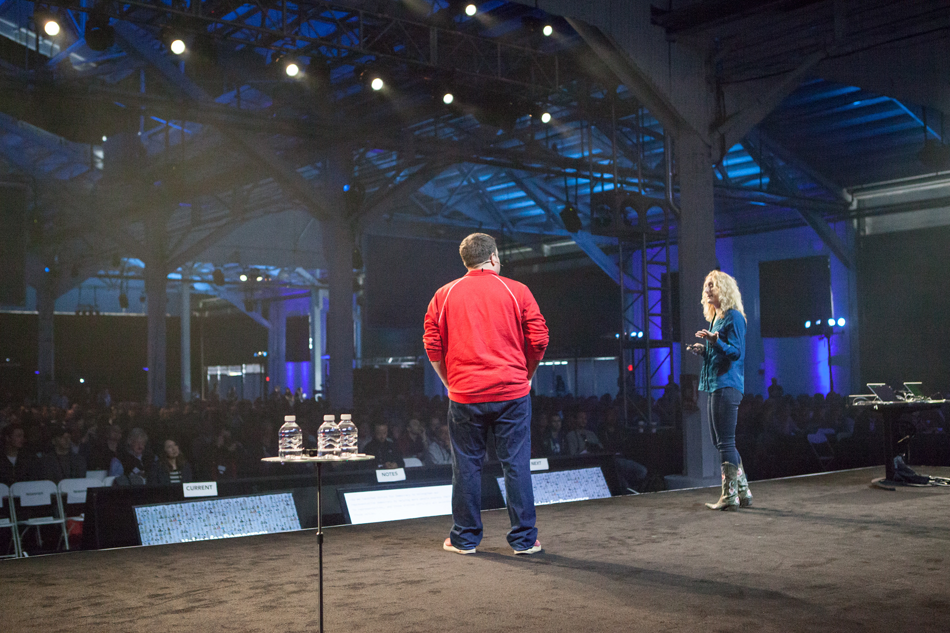 Two speakers on stage in industrial warehouse venue with blue uplighting